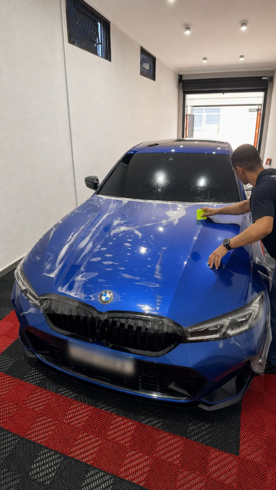 Profissional da MH instalando PPF transparente no capô de uma BMW azul, com a placa desfocada